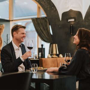 Fröhliches Gespräch eines Paares bei Wein und Essen im Restaurant Woodfire im Hotel Grauer Bär Innsbruck.