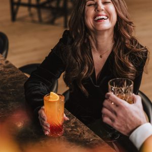 Fröhliche Frau mit Cocktailglas im stilvollen Licht des Restaurants Woodfire im Hotel Grauer Bär Innsbruck.